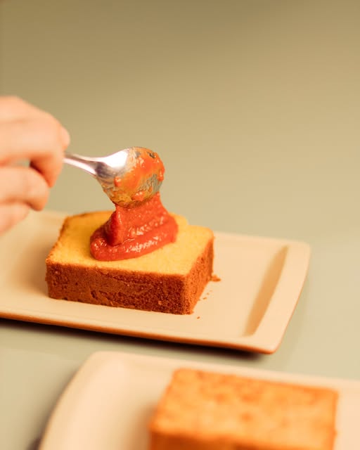 Fatia de bolo de fubá recebendo uma colherada de goiabada, em prato bege.